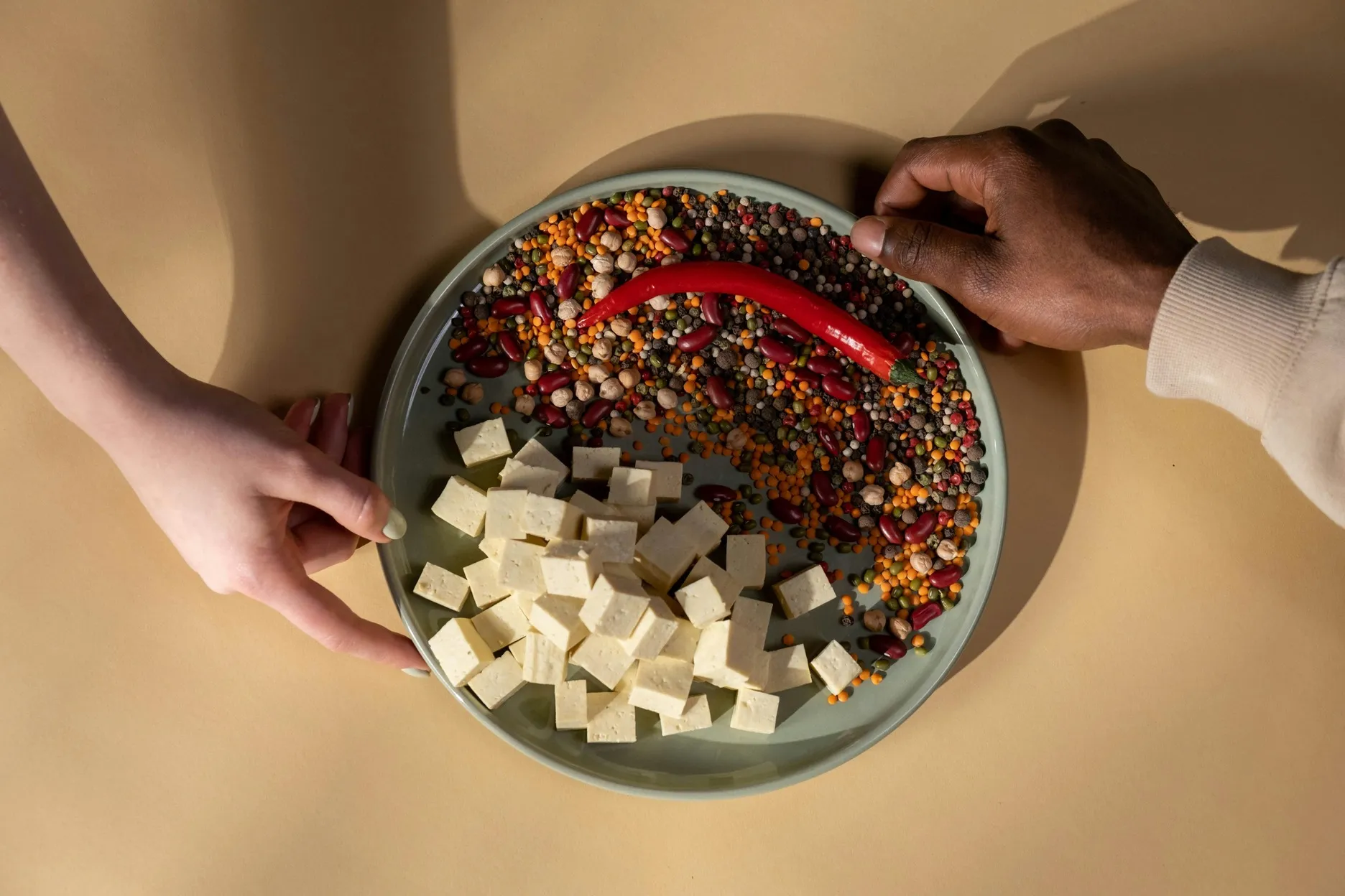 Plate with tofu cubes, mixed legumes, and a red chili pepper held by two hands on a beige background