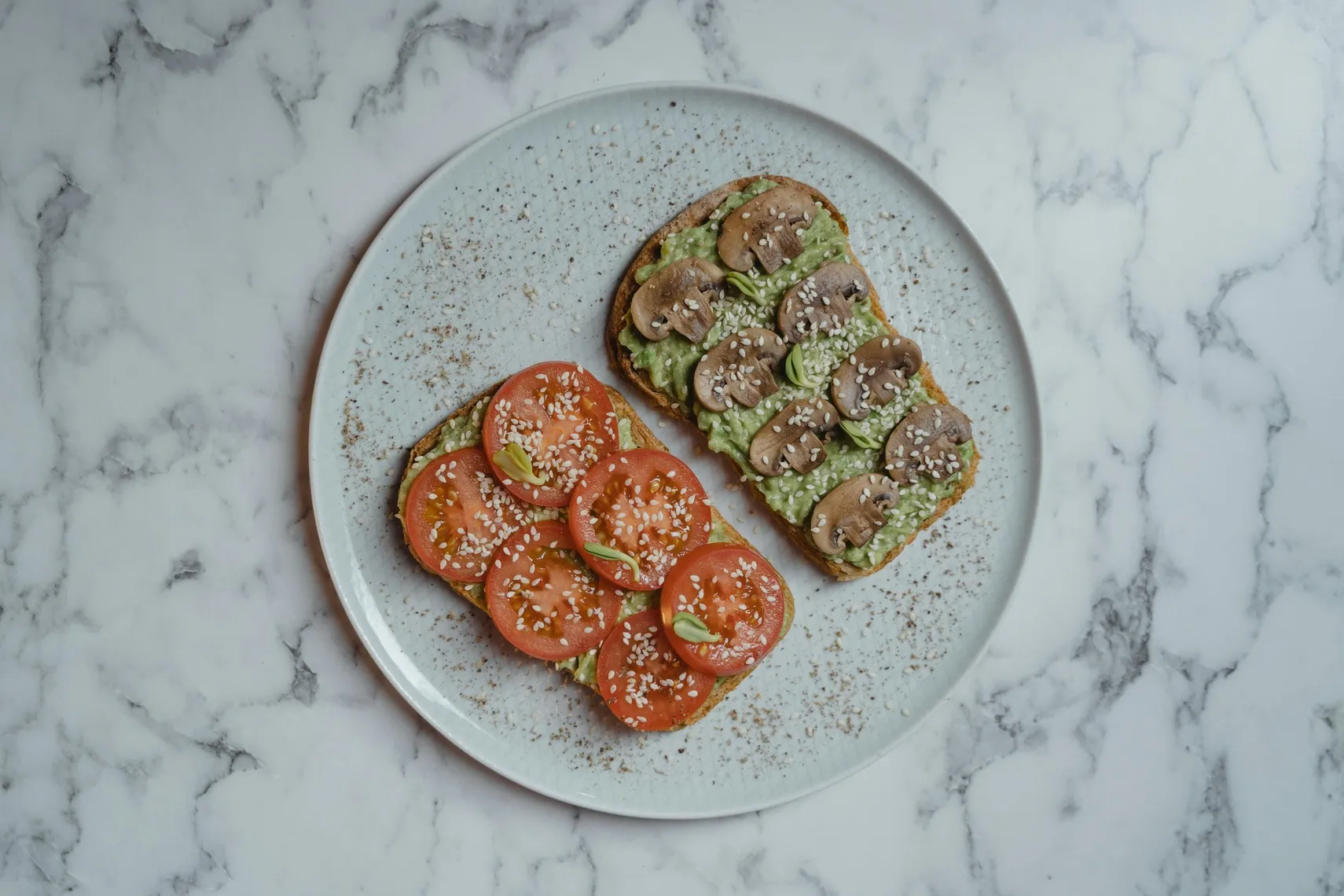 Two slices of avocado toast topped with tomatoes, mushrooms, and sesame seeds on a white plate